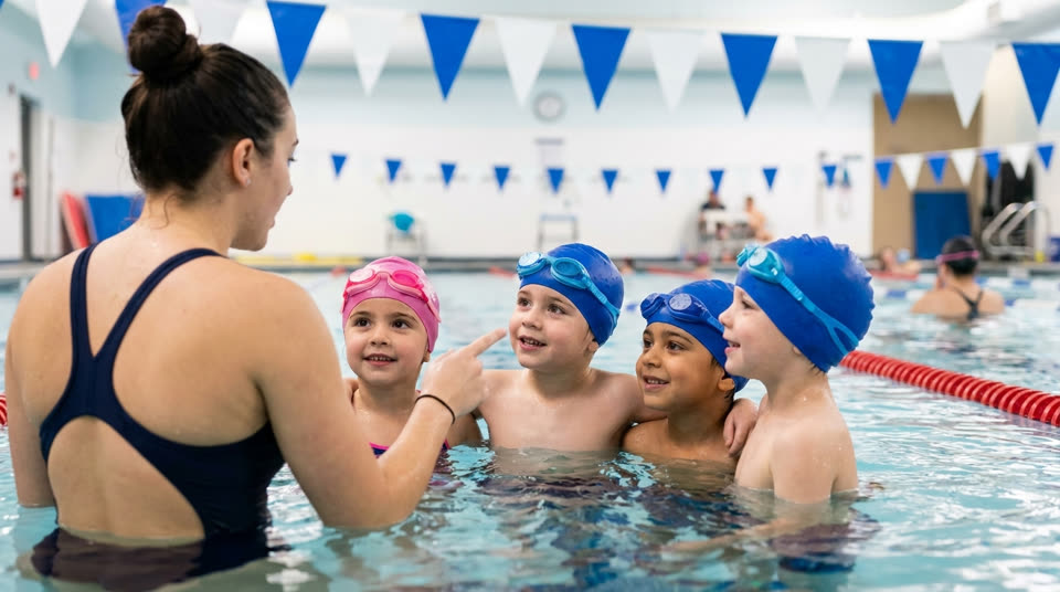 A group swim lesson: children waiting in a row at the pool edge while one instructor works with a single child